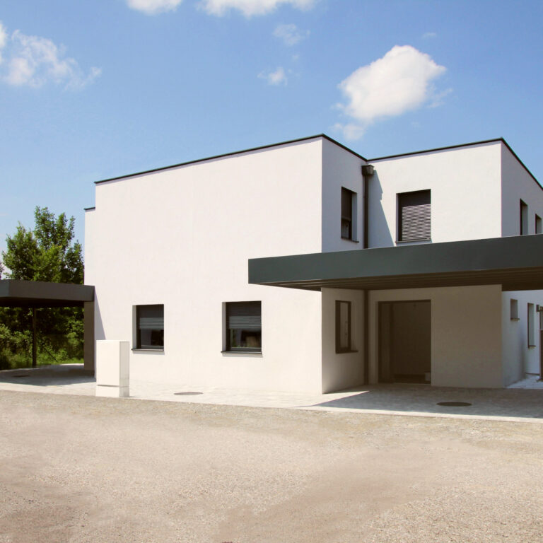 Completed duplex house with simplified carport and white façade, reflecting cost-saving adaptations.