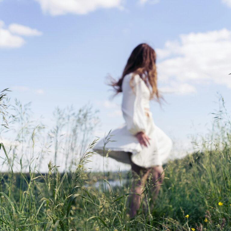 Woman in a white dress moving through a sunlit meadow, evoking calm, freedom and a slower pace of life.